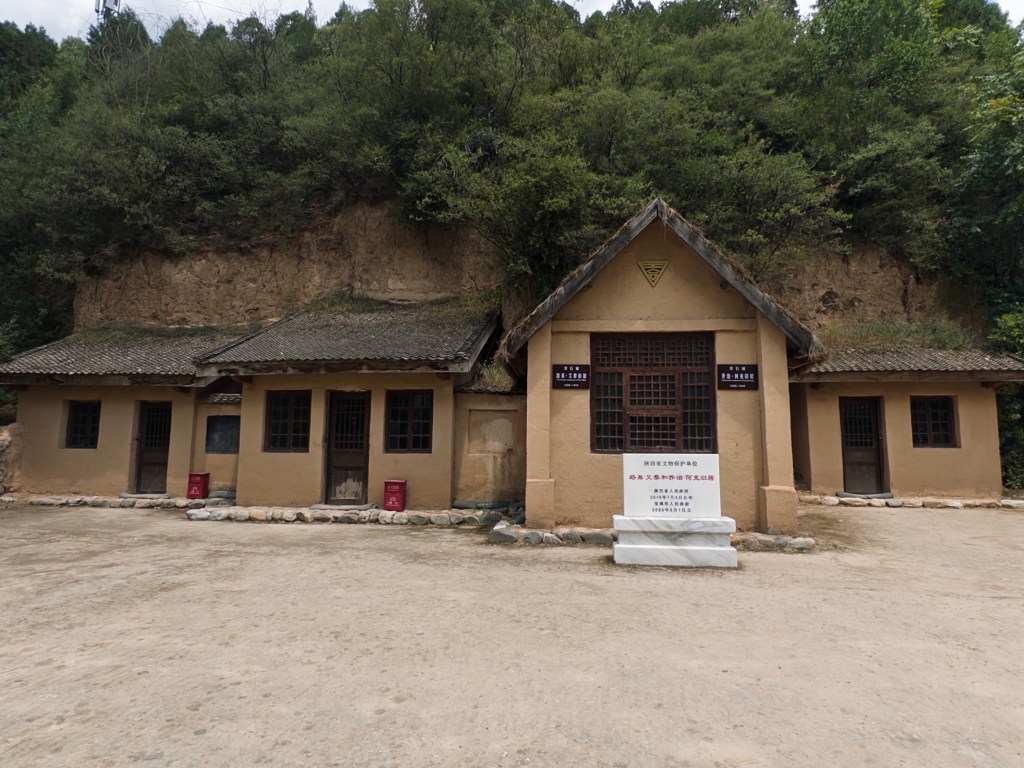 A historical building made of earth and wood, featuring a traditional sloped roof, with a sign in front and surrounded by greenery and steep terrain.