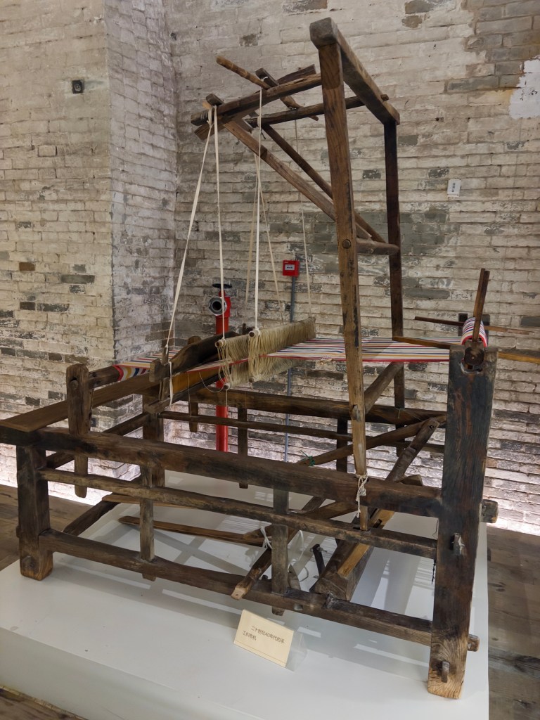 A vintage wooden loom displayed in a rustic setting, featuring a simple structure with horizontal and vertical wooden beams, strings, and colorful threads used for weaving.