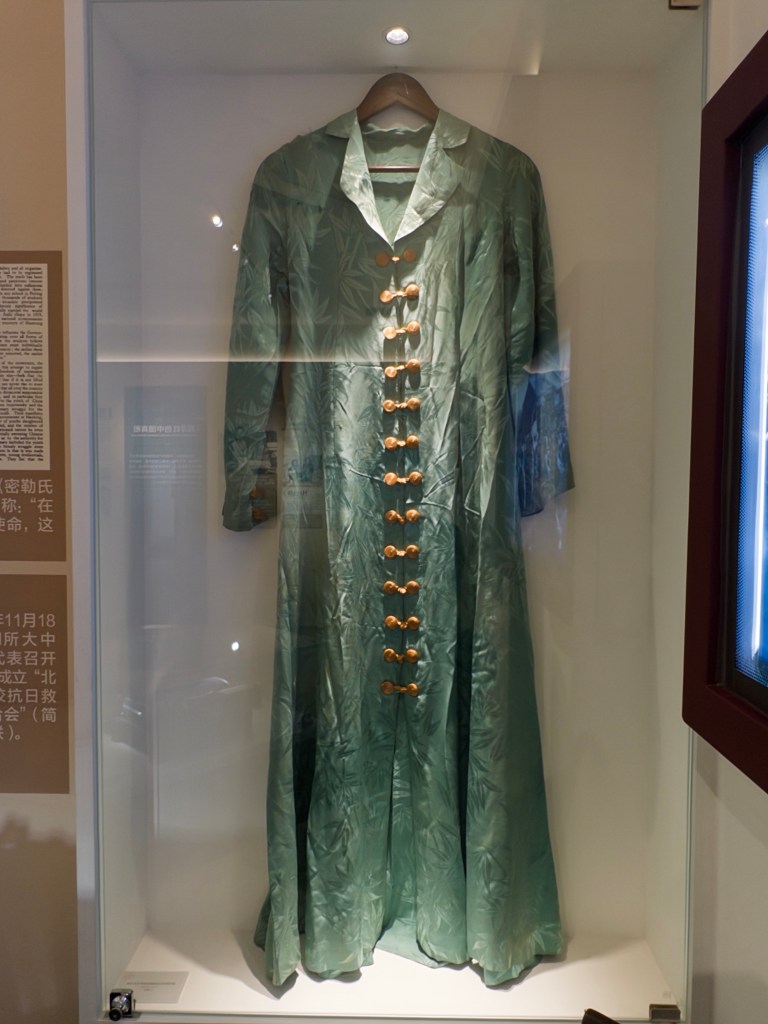 A vintage green silk coat with a collar and wooden buttons, displayed in a glass case.
